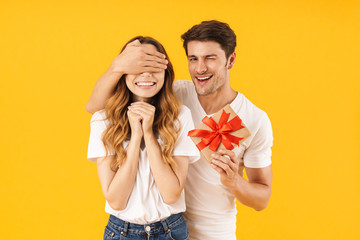 Portrait of happy couple in basic t-shirts standing together while man holding present box and covering woman's eyes