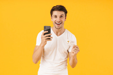 Poster - Photo of brunette man in basic t-shirt smiling at camera while holding smartphone and credit card