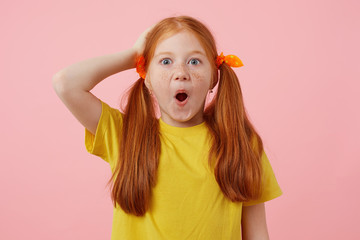 Wall Mural - Portrait of happy amazed petite freckles red-haired girl with two tails, wears in yellow t-shirt, stands over pink background with wide open mouth.