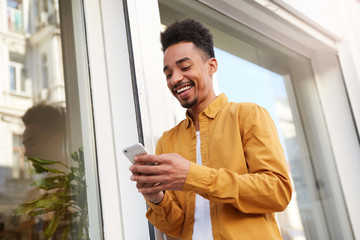 Wall Mural - Phortrait of young cheerful dark skinned guy in yellow shirt walking down the street , holds telephone, chatting with girlfriend, broadly smiling, looks happy.