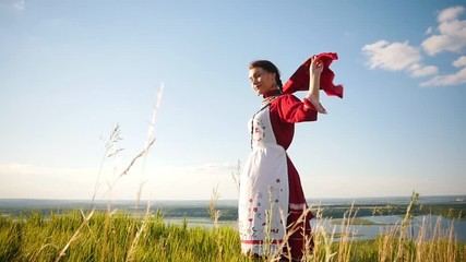 Wall Mural - A young woman in russian traditional clothes standing on the field - handkerchief flutters by the wind