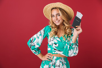 Wall Mural - Cheerful young blonde girl tourist in bright blue dress posing isolated over red wall background holding passport with tickets.