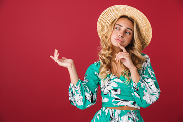 Wall Mural - Thinking thoughtful cute young blonde girl in bright blue dress posing isolated over red wall background looking aside wearing hat showing copyspace.