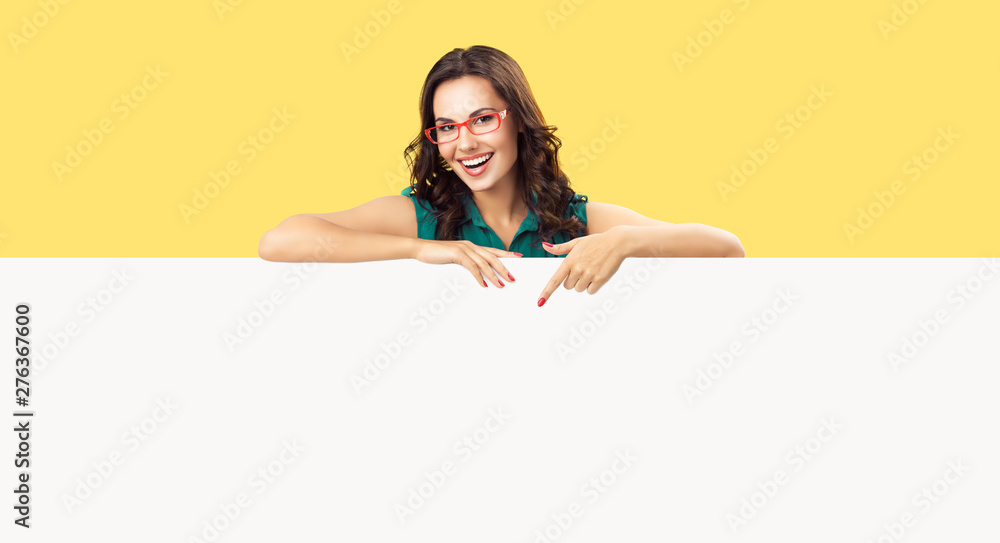 Happy smiling beautiful young woman in green clothing and red glasses, pointing blank white bill board or copy space empty place for ad slogan or sign text, over yellow color background - obrazy, fototapety, plakaty 