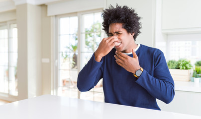 Poster - Young african american man wearing casual sweater sitting at home smelling something stinky and disgusting, intolerable smell, holding breath with fingers on nose. Bad smells concept.