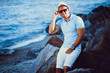 © zamuruev - The man at the resort in a white shirt and sunglasses, hat sitting on a rock on the sea background.