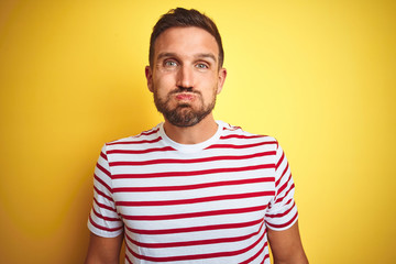 Poster - Young handsome man wearing casual red striped t-shirt over yellow isolated background puffing cheeks with funny face. Mouth inflated with air, crazy expression.