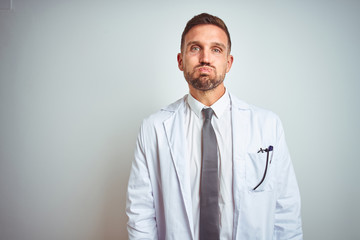 Poster - Young handsome doctor man wearing white profressional coat over isolated background puffing cheeks with funny face. Mouth inflated with air, crazy expression.