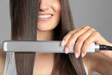 Wall Mural - Young woman using hair iron on grey background, closeup