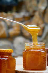 A table with a wicker table top, honey in honeycombs on a wooden board, honey flows from a spoon into an open glass jar. Medicinal herbs oregano, chamomile and strawberry flowers.