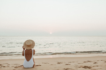 Summer vacation fashion concept. Young, tanned woman wearing a beautiful white swimsuit with a straw hat is sitting and relaxing on tropical beach with white sand and is watching sunset and sea.