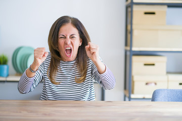 Middle age senior woman sitting at the table at home angry and mad raising fists frustrated and furious while shouting with anger. Rage and aggressive concept.