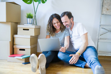 Middle age senior romantic couple in love sitting on the apartment floor with boxes around and using computer laptop smiling happy for moving to a new home