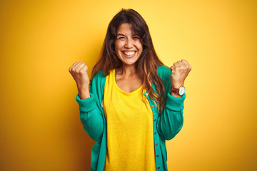 Wall Mural - Young woman wearing t-shirt and green sweater standing over yelllow isolated background celebrating surprised and amazed for success with arms raised and open eyes. Winner concept.