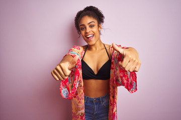 American woman on holiday wearing bikini and colorful dress over isolated pink background approving doing positive gesture with hand, thumbs up smiling and happy for success. Winner gesture.