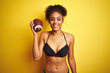 © Krakenimages.com - Afro american woman on vacation wearing bikini playing rugby over isolated yellow background with a happy face standing and smiling with a confident smile showing teeth