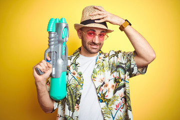 Sticker - Young man wearing hawaiian flowers shirt holding water gun over yellow isolated background stressed with hand on head, shocked with shame and surprise face, angry and frustrated.