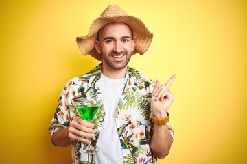 Wall Mural - Young man wearing summer hawaiian flowers shirt and drinking a cocktail over yellow background very happy pointing with hand and finger to the side