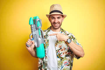 Wall Mural - Young man wearing hawaiian flowers shirt holding water gun over yellow isolated background very happy pointing with hand and finger