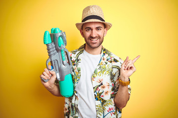 Sticker - Young man wearing hawaiian flowers shirt holding water gun over yellow isolated background very happy pointing with hand and finger to the side