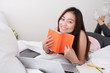 © weedezign - Asian woman wear pink tank top lying on bed reading book with research job on laptop in bedroom.work at home.e-larning concept.study online.