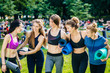 © Iryna - Team of five fitness women hugging, laughing talking in park in summer morning. Females posing over big group of attending a yoga class on background. Excited emotions sport recreations concept.