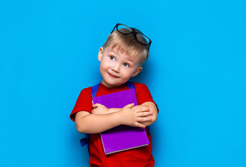 small happy smiling boy with glasses on his head, book in hands, schoolbag on his shoulders. back to