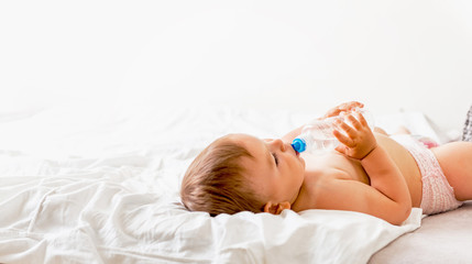 Wall Mural - Baby toddler sits on the white bed, smiles and drinks water from plastic bottle. Copy space