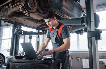 Half of the job is done. Man at the workshop in uniform using laptop for his job for fixing broken car