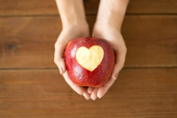 food, valentines day and health concept - close up of hands holding ripe red apple with carved heart shape over wooden table