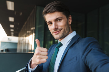 Photo of successful businessman showing thumb up and taking selfie while standing outside job center