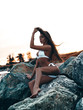 © maksimvostrikov - Beautiful, sexy young girl with an attractive slim shapely body in bikini is sitting and posing on the rocks at the sunset. Summer hot outdoor photoshoot on the shore. Photo on the beach