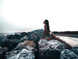 © maksimvostrikov - Elegant, sexy young woman with a tempting slim shapely body in bikini is sitting and posing on the rocks at the sunset. Summer outdoor photoshoot on the shore. Photo on the beach near the water