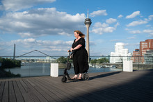 Overweight Woman Wearing Black Dress Riding An Electric Scooter On A Bridge In Front Of Modern Architecture Cityscape At Medienhafen Duesseldorf On A Sunny Summer Day