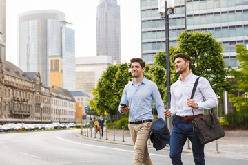 Smiling young two friends colleagues business men walking outdoors looking aside holding phone.