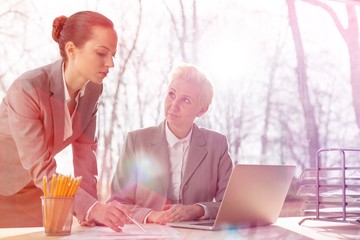 Wall Mural - Confident mature businesswoman looking at colleague explaining document at desk against window in office