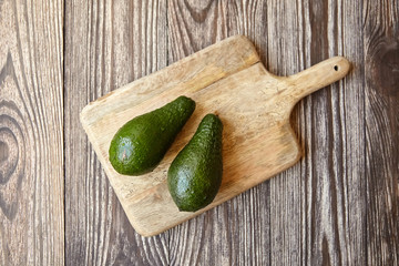 Avocados on a natural pine cutting board on wooden background. Two whole green fresh tropical fruits on brown table, top view