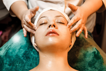 Close up portrait of a amazing caucasian woman having a facial massage by a female hands in a wellness spa center while leaning on a bed with closed eyes.