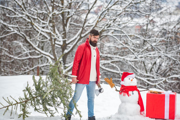 Canvas Print - Bearded man with snowman is carrying Christmas tree in the wood. A handsome young man with snow man carries a Christmas tree. Merry Christmas and happy new year greeting card. Snowman with gift.