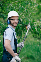 cheerful gardener in helmet, protective glasses and noise-canceling headphones holding telescopic pole saw and smiling at camera