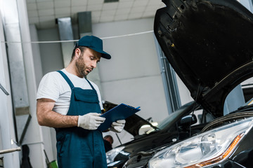 handsome bearded repairman in cap holding pen and clipboard near car