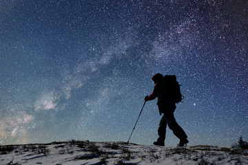silhouette of male tourist with backpack and trekking sticks going along snow-covered path high up i