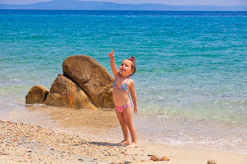 Cute little girl points in the sky at the beach near the sea on vacations.