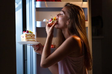 Hungry woman in pajamas eating sweet cakes at night near refrigerator. Stop diet and gain extra pounds due to high carbs food and unhealthy night eating