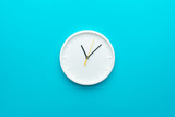 White wall clock with yellow second hand hanging on the wall. Minimalist flat lay image of plastic wall clock over blue turquiose background with copy space and central composition.