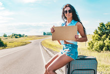 Pretty Young Woman Hitchhiking Along A Road And Waiting On A Country Road With Her Suitcase, Empty Cardboard Plate. Beautiful Girl In Sunglasses Hitchhiker. Empty Board