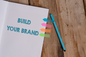 Conceptual hand writing showing Build Your Brand. Concept meaning enhancing brand equity using advertising campaigns Striped notebook colored arrow banners ballpoint wooden background