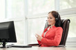 © spaskov - Young attractive woman in red jacket sitting on desk at head of executive in red jacket. Holds video conference in headphones with microphone. Drinking coffee from white cup.