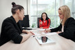 © spaskov - Three young attractive women in business suits are sitting at a desk and discussing workflows. Head and subordinates. Working team of professionals and colleagues.