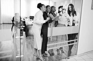 Wall Mural - Business womans pointing on glass with colorful paper notes. Diverse group of female employees in formal wear using stickers.